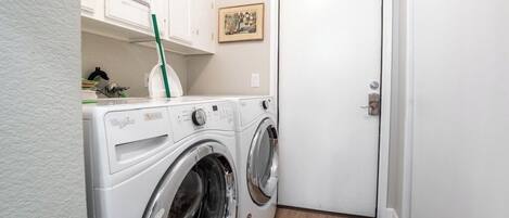 Een kleine wasruimte met een voorlader wasmachine en droger, een klein kastje boven de wasmachine, en een deur aan de rechterkant.