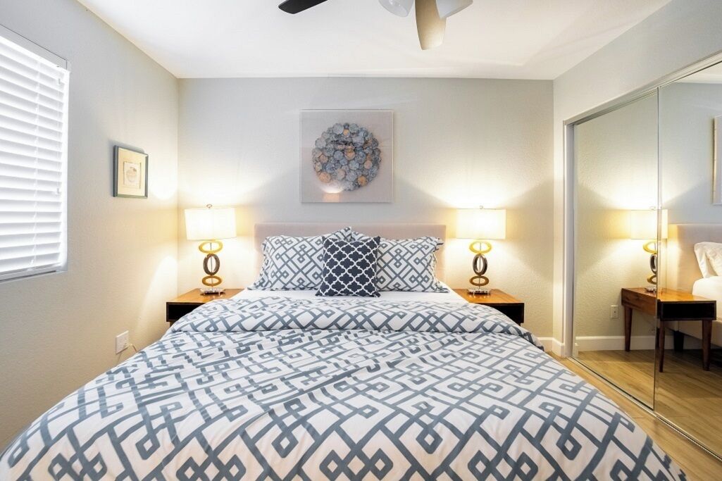 Un dormitorio con cama, dos mesitas de noche, ventilador de techo, un cuadro en la pared y espejo.