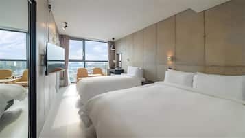 A hotel room with two beds, a desk, a chair, and a large window offering a city view.