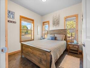 Une chambre à coucher avec un lit en bois, deux fenêtres équipées de stores, une table de chevet avec une lampe et une œuvre d’art accrochée au mur.
