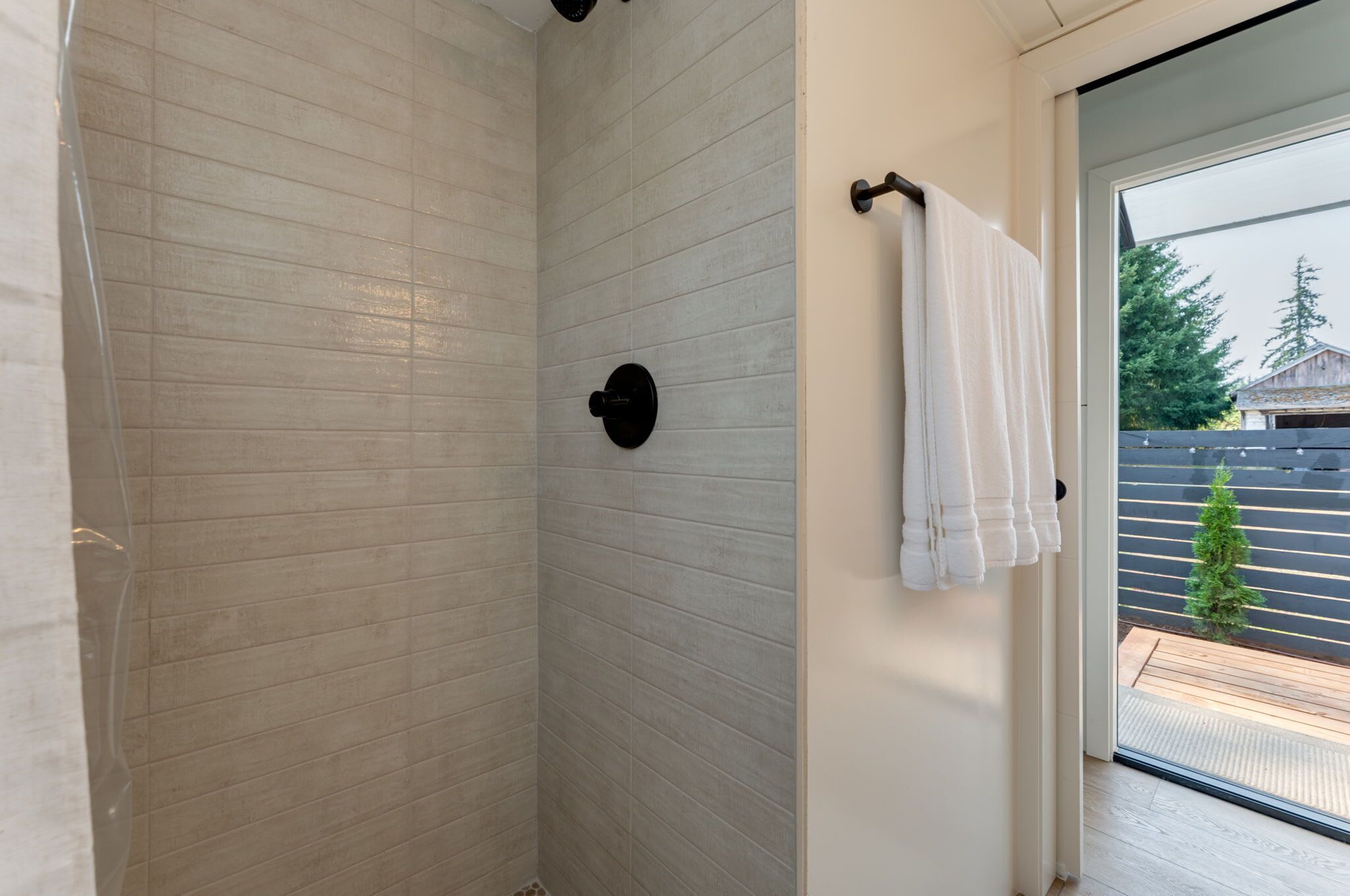 A shower with a tiled wall, a towel rack with a towel, and a view of a wooden deck and trees outside.