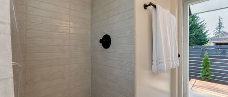 A shower with a tiled wall, a towel rack with a towel, and a view of a wooden deck and trees outside.