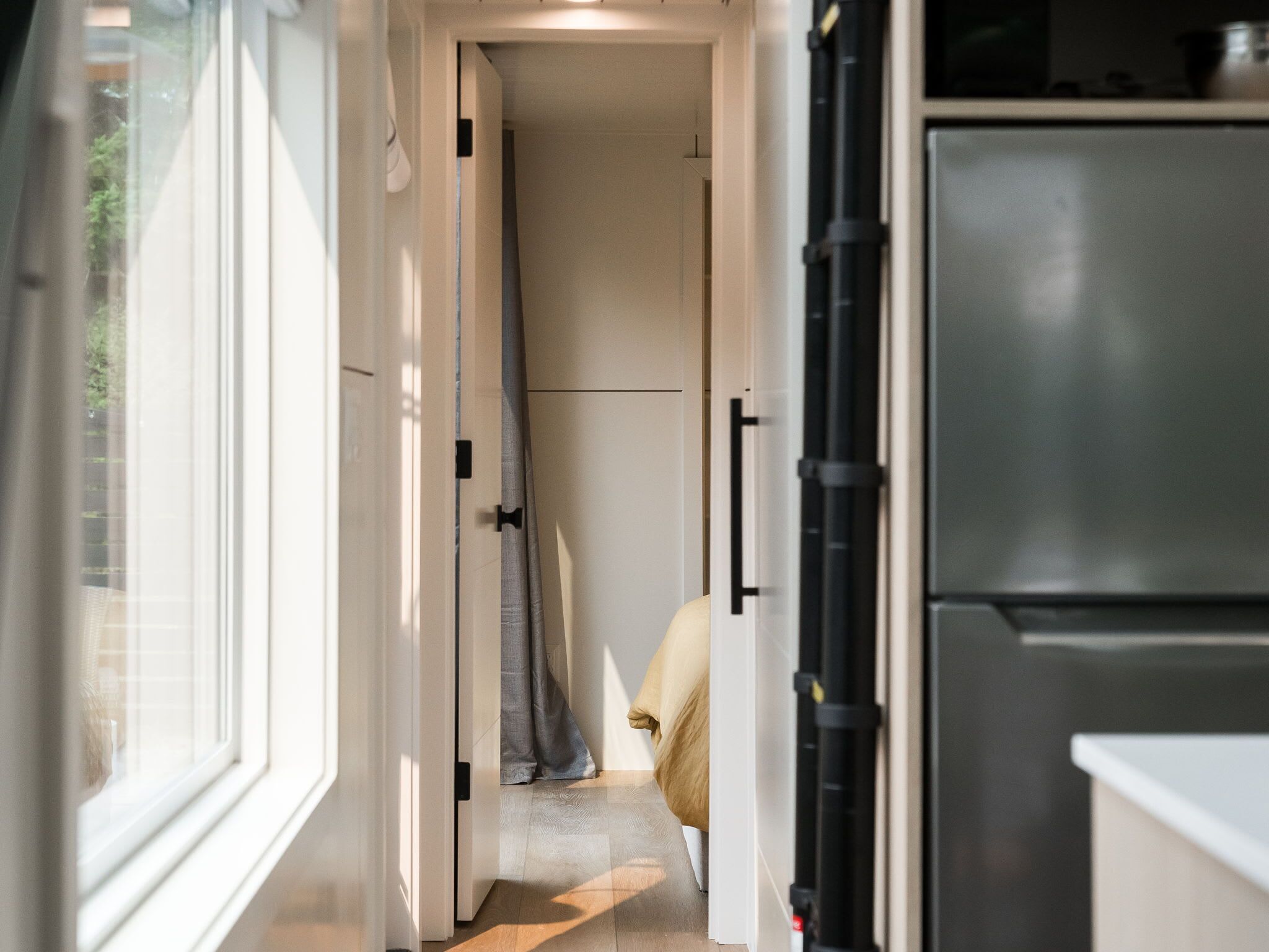 Un pasillo estrecho con una puerta que da a una habitación, un refrigerador a la derecha y una ventana a la izquierda.
