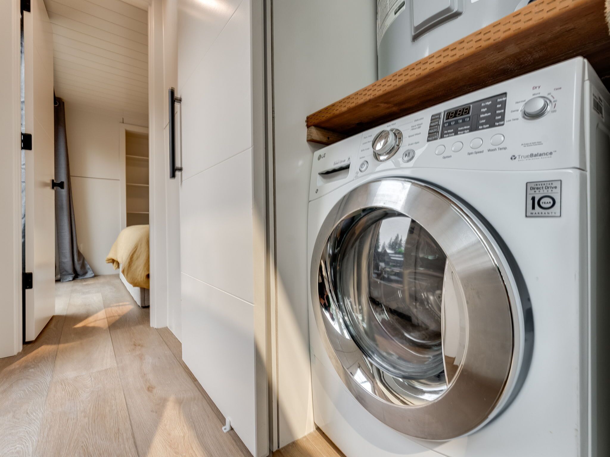 A modern laundry room with a front-loading washing machine, a wooden shelf above the machine, and a hallway leading to a closet.