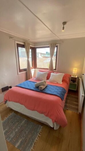 A bedroom with a bed, a nightstand, a lamp, and a view through the window. - Patagon Backpackers (Coyhaique)