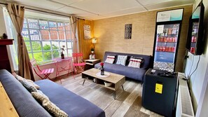 A furnished living room with a sofa, a small table, a chair, a refrigerator, and a window with curtains. - Patagon Backpackers (Coyhaique)