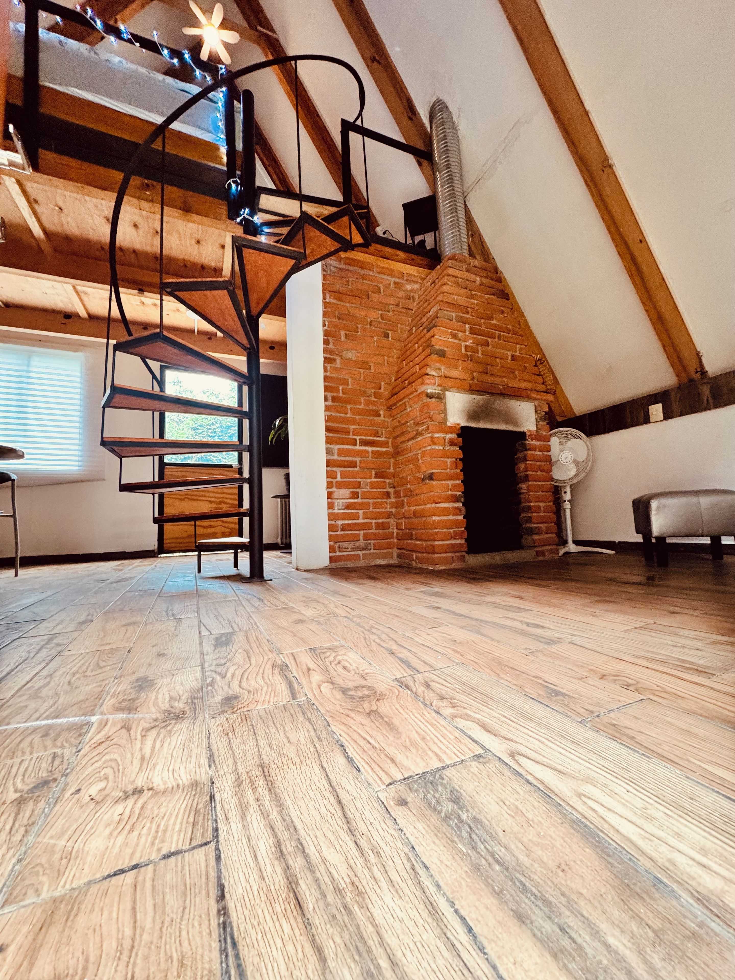 A loft-style room with a brick fireplace, wooden beams, a spiral staircase, and a large window.