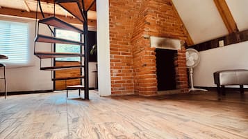 A loft-style room with a brick fireplace, wooden beams, a spiral staircase, and a large window.