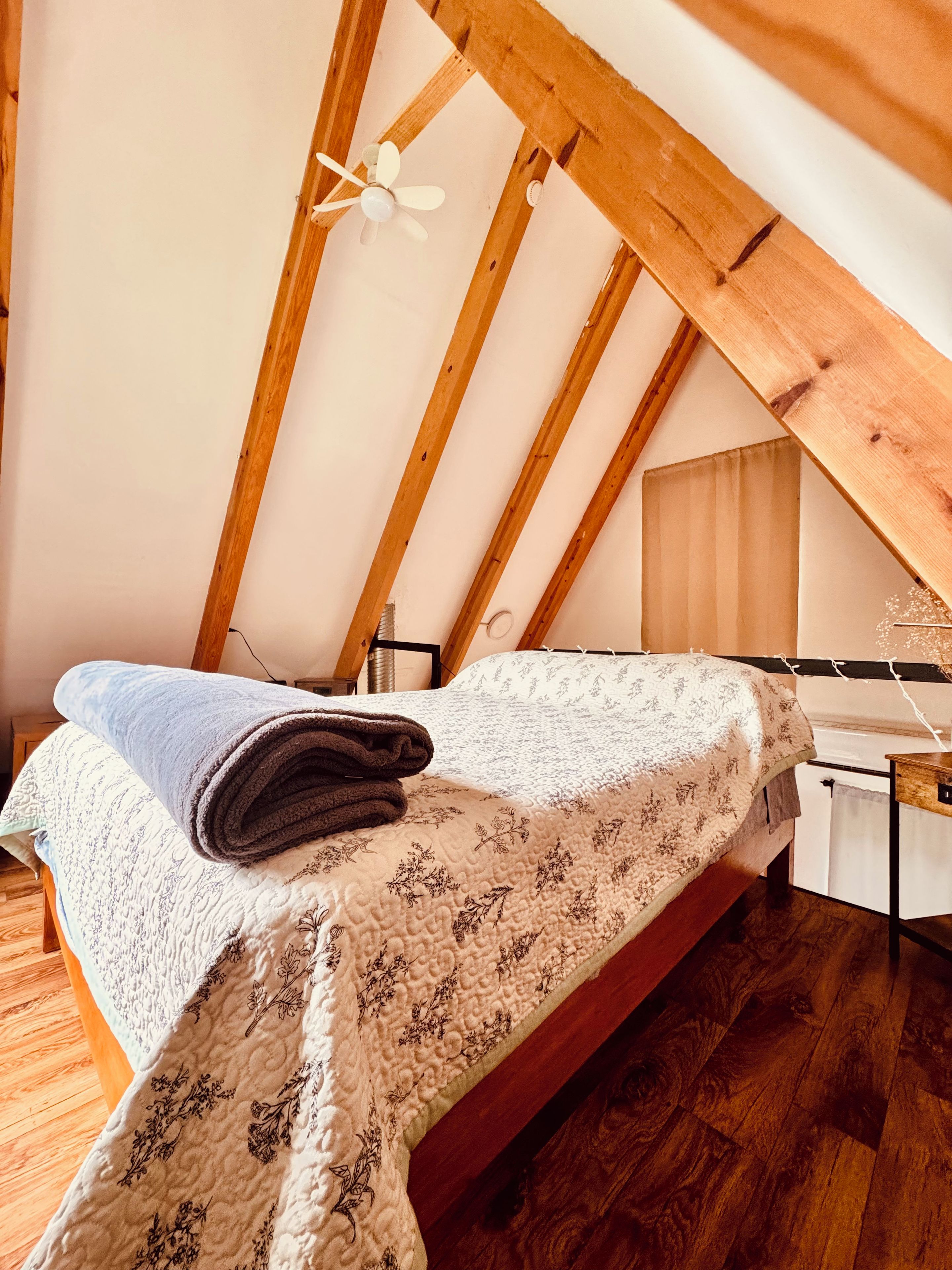 Une chambre avec un plafond en pente, des poutres en bois, un lit recouvert d’une couvre-lit à motifs floraux, un bureau et une chaise.
