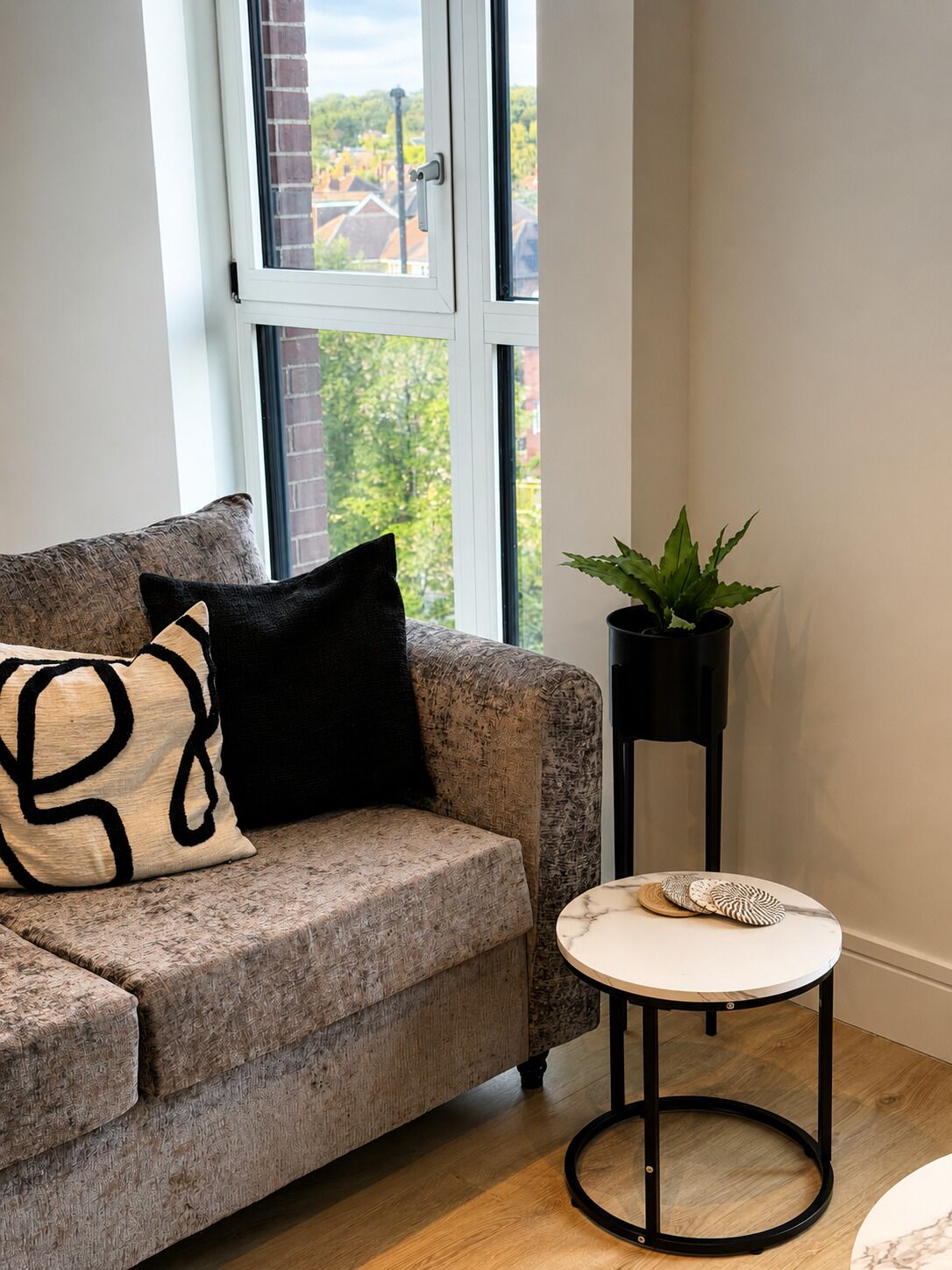 A living room with a sofa, a small round table, a potted plant, and a window with a view of buildings and trees.