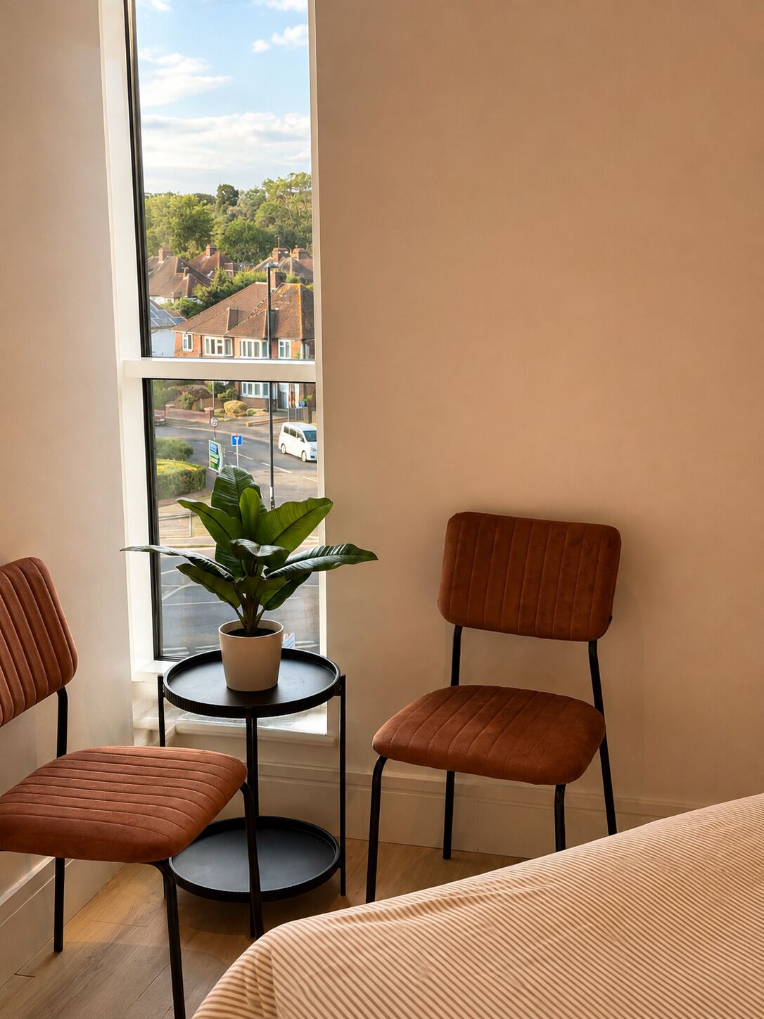 A room with a large window, two chairs, a small table with a plant, and a bed.