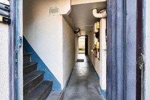 Un pasillo estrecho con una escalera a la izquierda que lleva a una puerta. El pasillo tiene piso de concreto y paredes pintadas en dos colores.