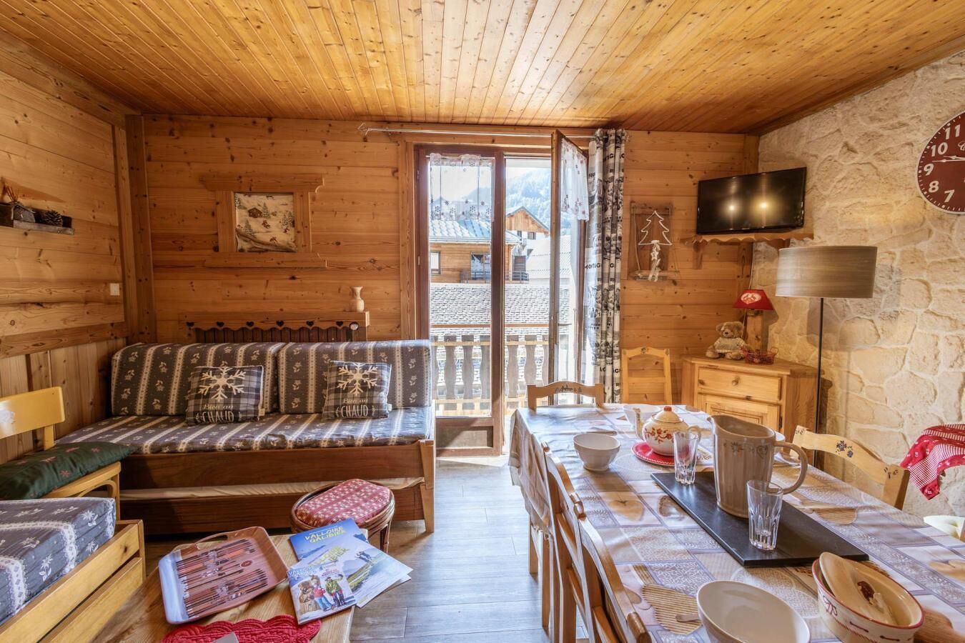 Una cabaña acogedora con interior de madera, un sofá con cojines estampados, una mesa de comedor con mantel rojo y una pared de piedra con un reloj.