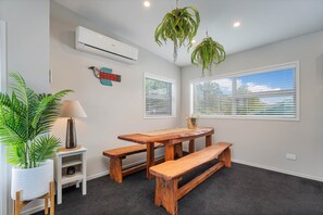 A room with a wooden table and benches, a white air conditioning unit, a potted plant, and a wall-mounted decorative piece. - Wharf Landing: The Apartment (Coromandel)