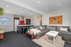A modern living room with a grey sofa, a coffee table, and a kitchen area in the background. - Wharf Landing: The Apartment (Coromandel)
