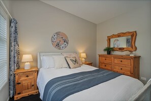 A bedroom with a bed, two bedside tables, a dresser, a mirror, and a wall decoration. - Wharf Landing: The Apartment (Coromandel)