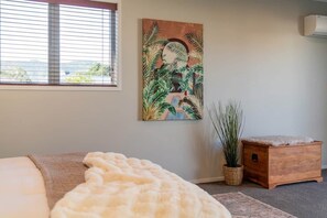 A bedroom with a bed, a large window, a painting, a plant, and a wooden chest. - Wharf Landing: The Studio (Coromandel)
