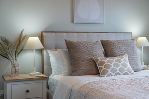 A neatly made bed with a headboard, two bedside lamps, and a bedside table with a vase of dried plants.