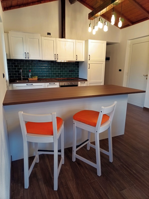 A kitchen with white cabinets, a dark countertop, and a green backsplash. There are two white bar stools with orange cushions.