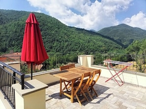 A terrace with a wooden table and chairs, a red umbrella, and a view of a mountainous landscape.