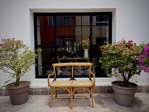 Un banco de madera con asiento tejido, flanqueado por dos macetas con follaje verde y flores de colores, ubicado frente a un ventanal con marco negro.