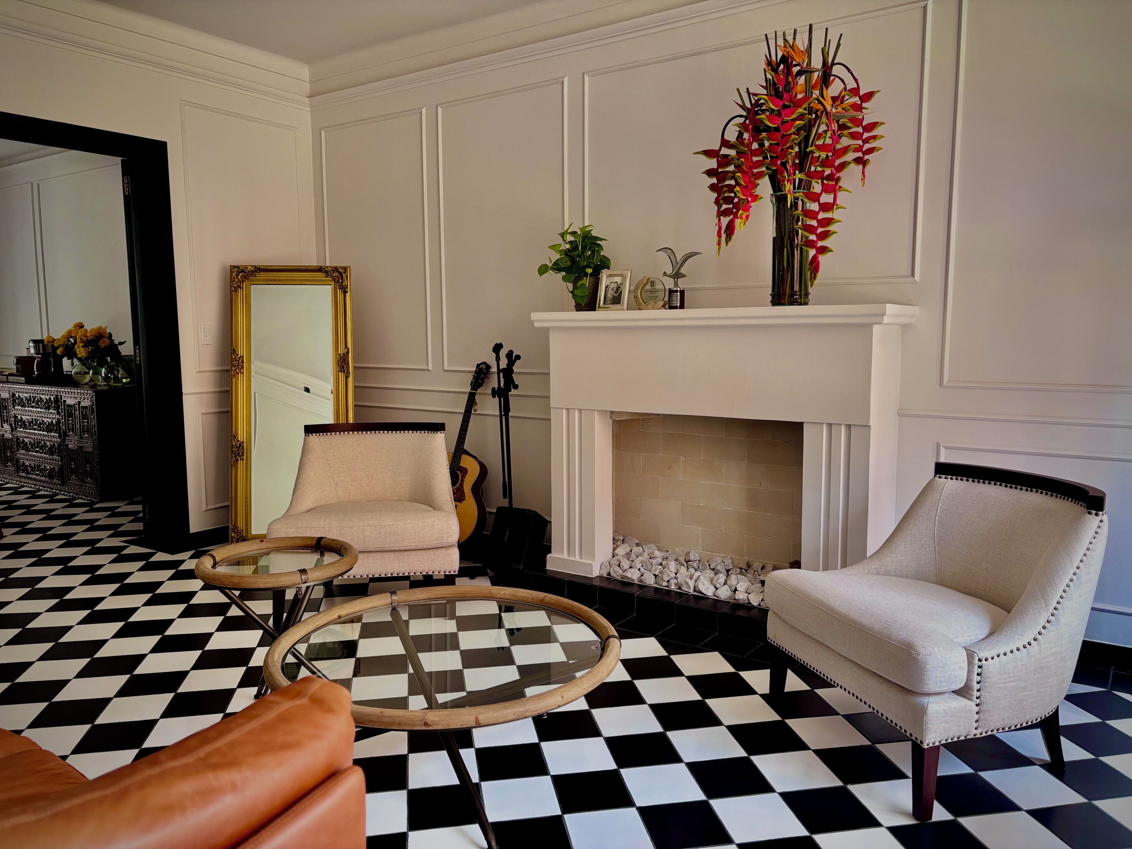 Una habitación con piso a cuadros, chimenea, dos sillones, un espejo y una guitarra.