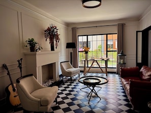 Una habitación con chimenea, suelo a cuadros, sofá de cuero, una silla, una guitarra, una lámpara y vistas al exterior.