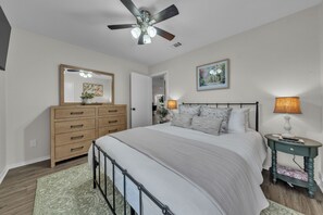 A bedroom with a bed, dresser, nightstand, lamp, ceiling fan, and artwork.