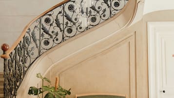 Un magnifique escalier orné de rampes en métal ouvragé, un banc recouvert d’un tissu vert et un lustre suspendu.