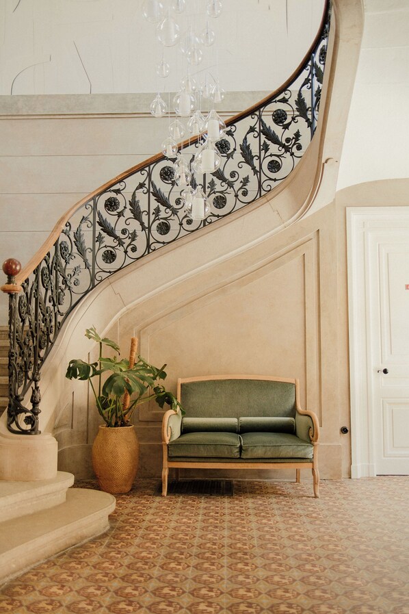 A grand staircase with ornate metal railings, a green upholstered bench, and a hanging chandelier.