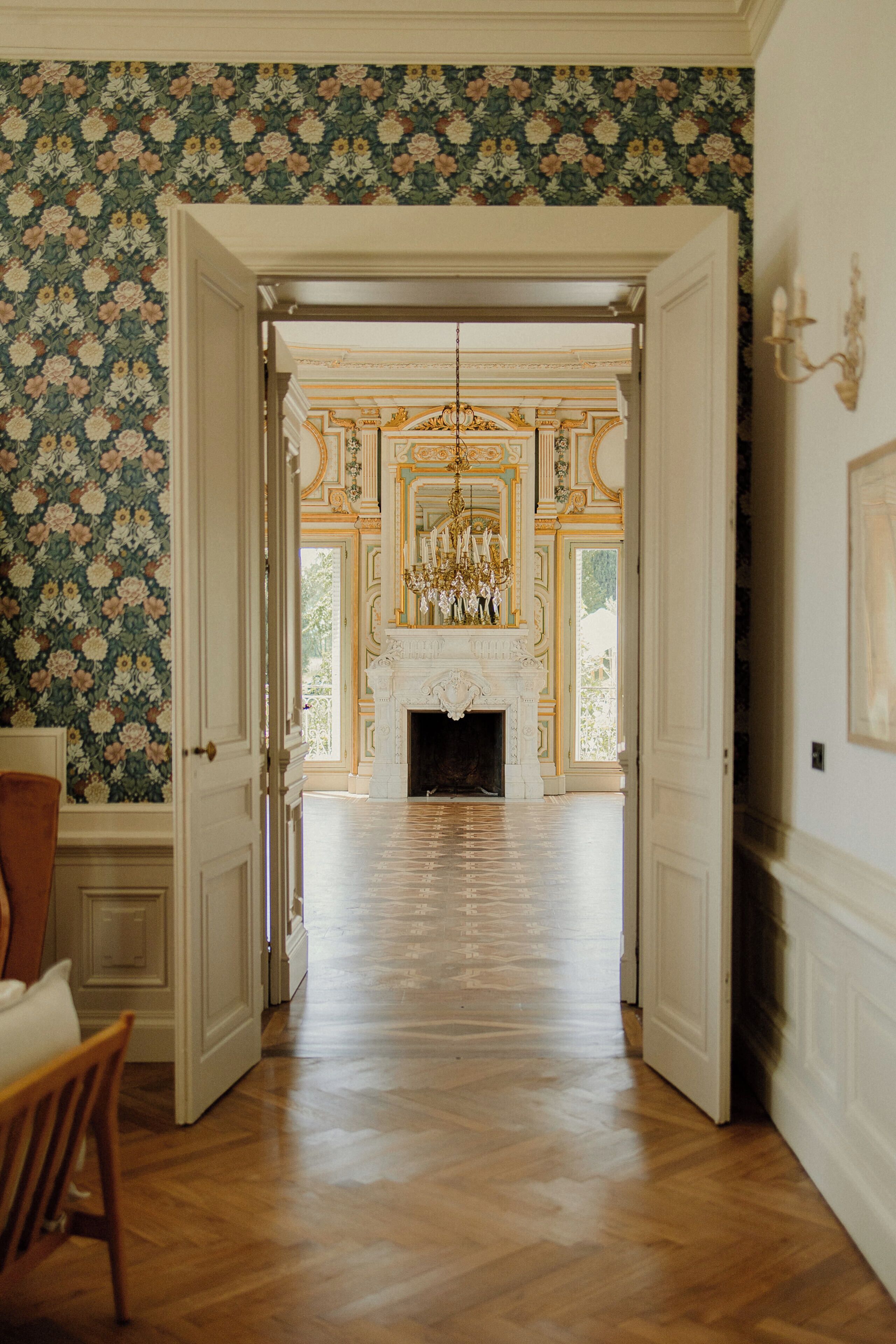 Un couloir mène à une vaste pièce dotée d’une cheminée, d’un plafond orné et d’un papier peint à motifs floraux.
