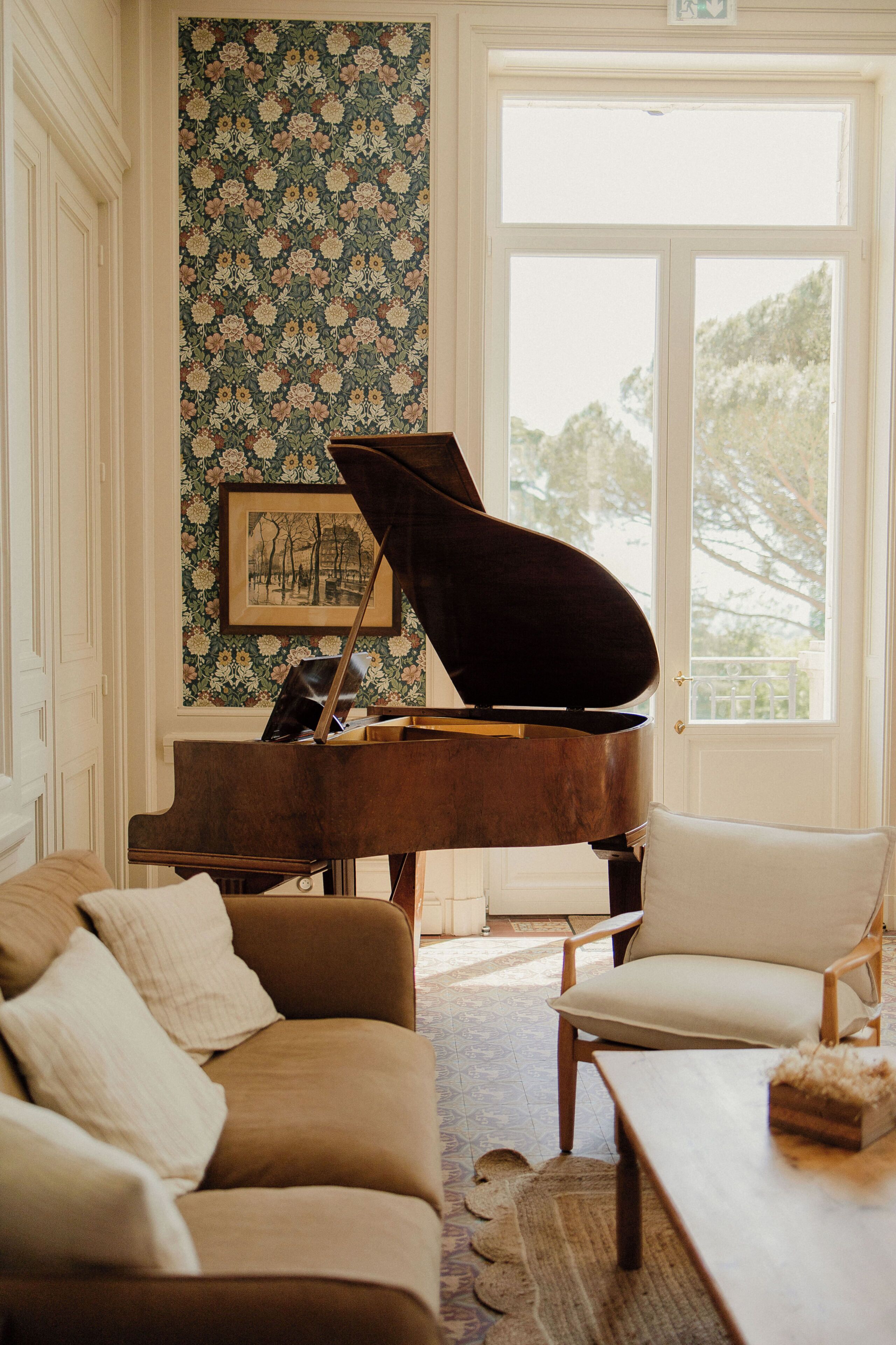 A grand piano in a well-lit room with a floral wallpaper, a framed picture on the wall, and a large window.