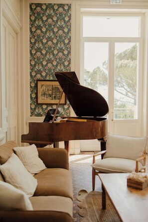 A grand piano in a well-lit room with a floral wallpaper, a framed picture on the wall, and a large window. - Château du Bost, renovated, heated pool, Beaujolais (Blacé)