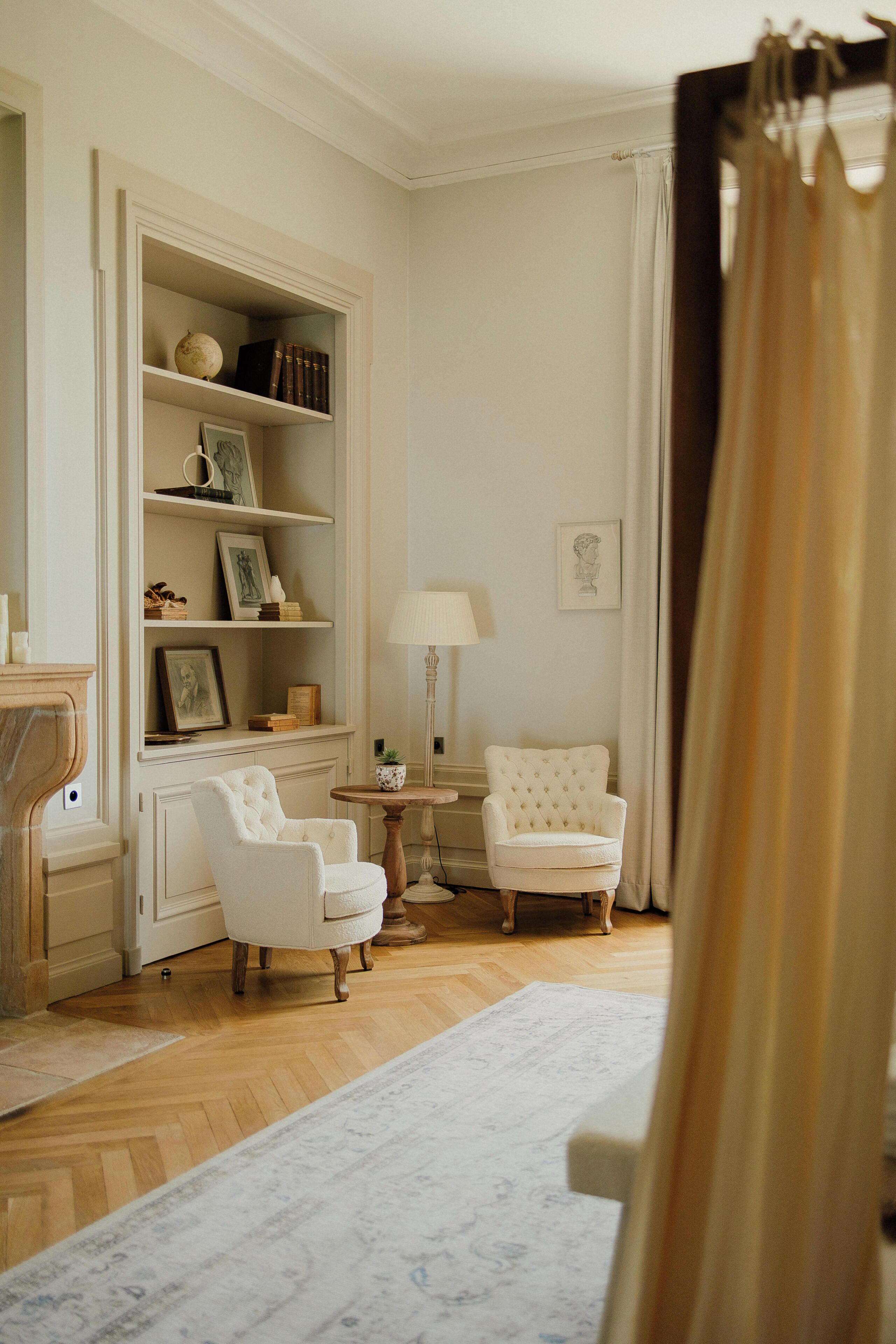 Una habitación con piso de madera, un sillón blanco, una pequeña mesa redonda con una lámpara, una estantería con libros y objetos decorativos, y un cuadro en la pared.