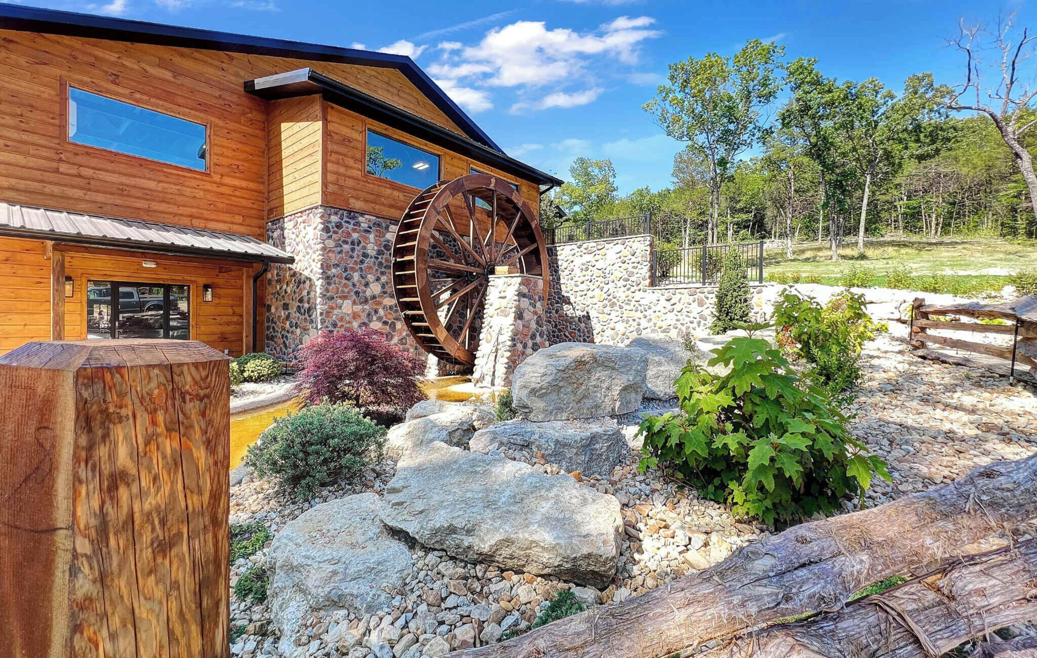 Un bâtiment en bois doté d’une roue à eau, entouré de rochers et de verdure.