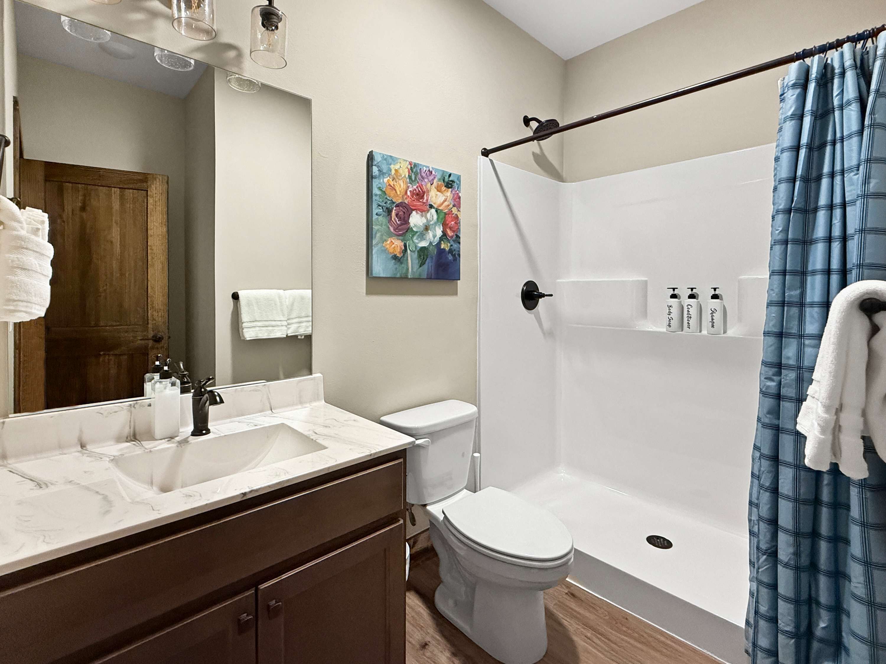 Une salle de bain avec un lavabo blanc, des toilettes, une douche avec un rideau bleu et un tableau accroché au mur.