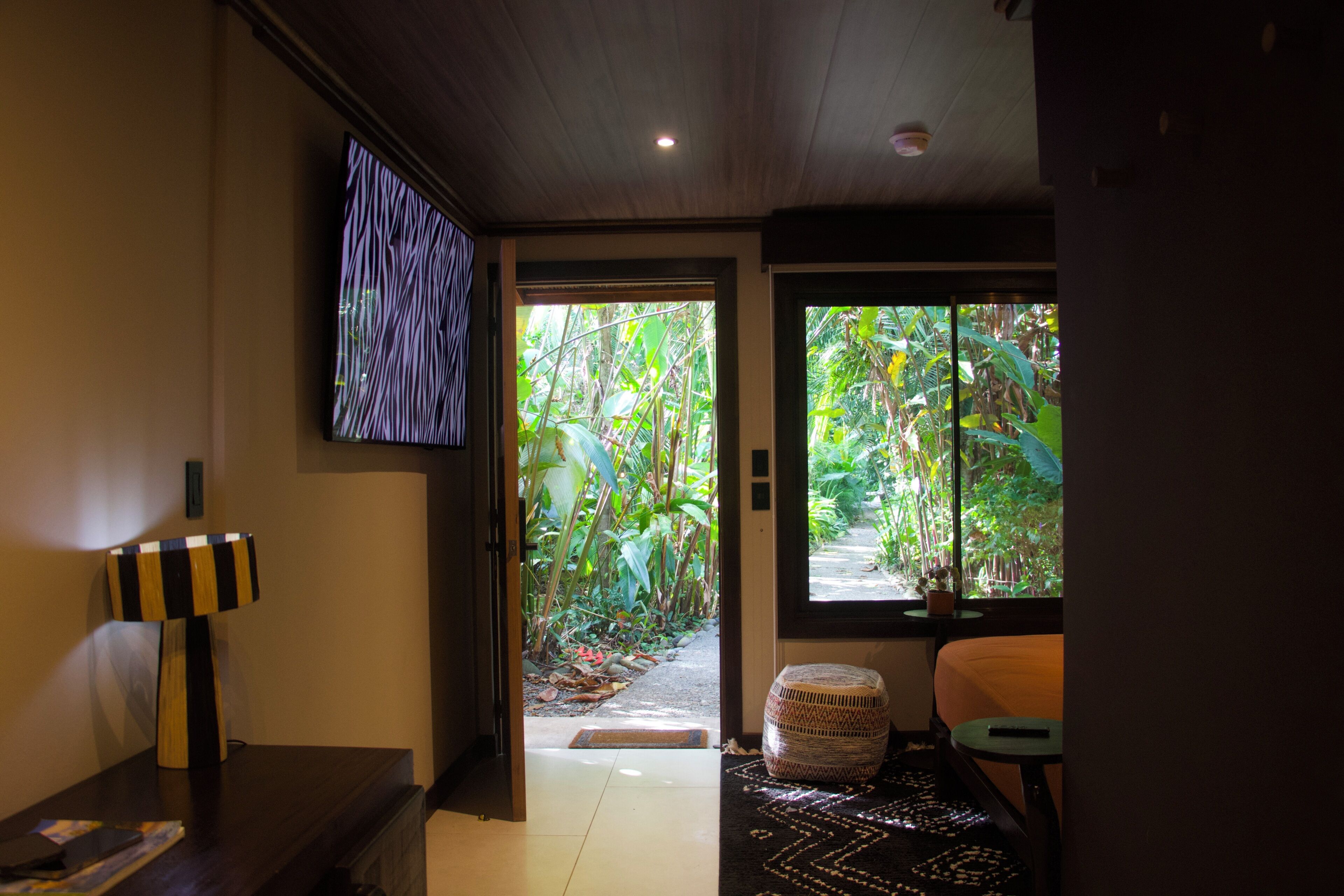 A hotel room with a television on the wall, a door leading to a garden, and a small table with a cushion.