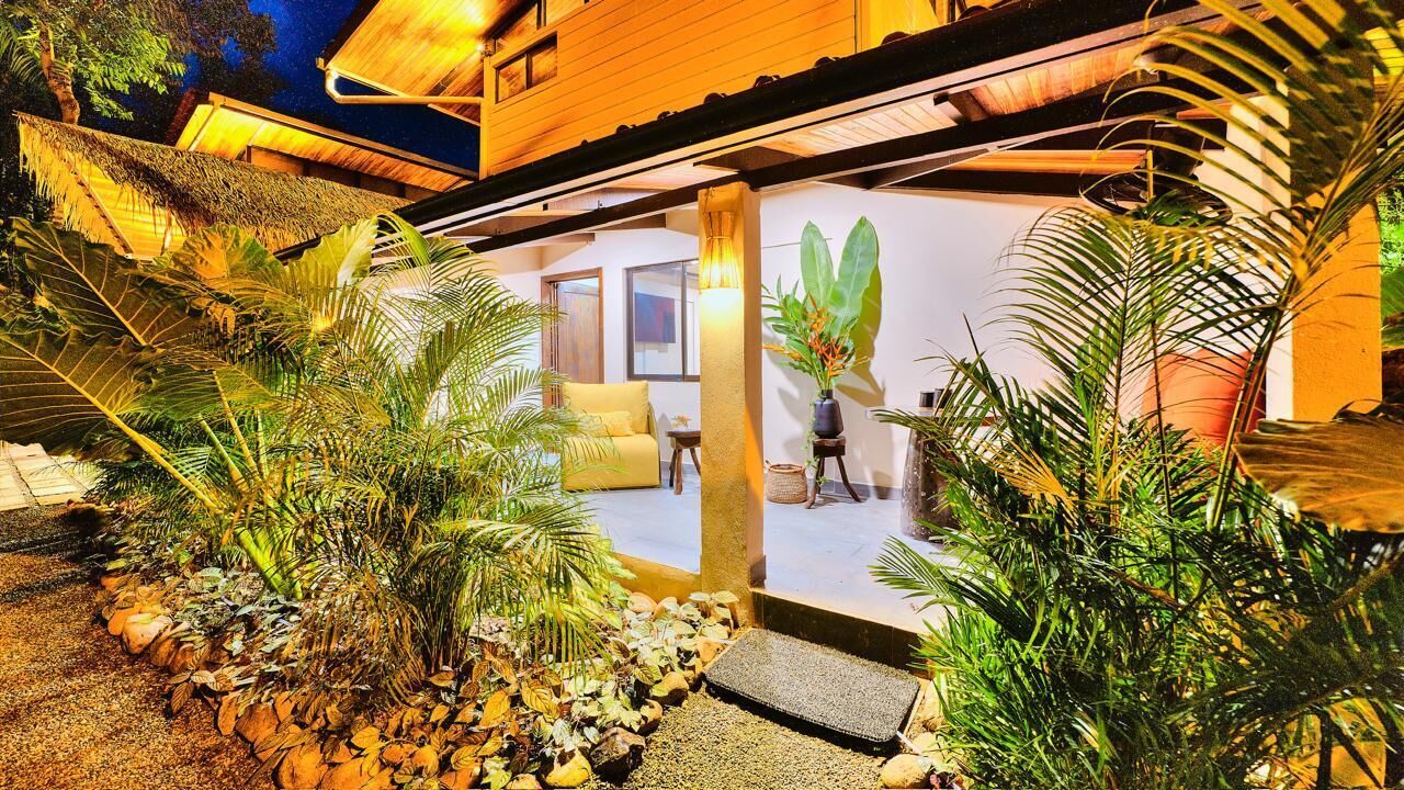 Un bâtiment moderne doté d’un toit de chaume, d’un jardin bien entretenu avec des plantes tropicales, et d’un intérieur visible où l’on aperçoit un canapé et une table.
