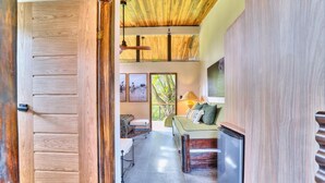 A room with a wooden door, a bed with pillows, a sofa, a lamp, and a window with a view of greenery. - Deluxe Jungle View Suite (4 twin) (Puerto Viejo)