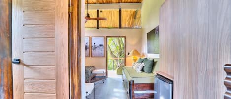 A room with a wooden door, a bed with pillows, a sofa, a lamp, and a window with a view of greenery.