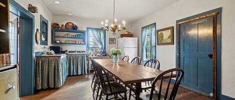 A dining area with a wooden table and chairs, a chandelier, and a kitchen area with shelves and a refrigerator.