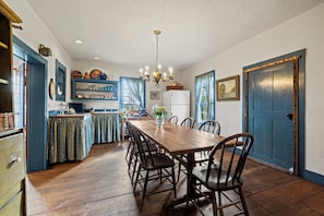 A dining area with a wooden table and chairs, a chandelier, and a kitchen area with shelves and a refrigerator. - A 1787 home older than the country it stands in (Fredericksburg)