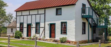 A two-story white building with a red roof, green window frames, and a wooden fence in front.