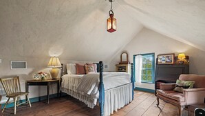 Ein Schlafzimmer mit einem hölzernen Bett, einem Sessel, einem Tisch mit Lampe und einem Fenster mit Vorhängen.