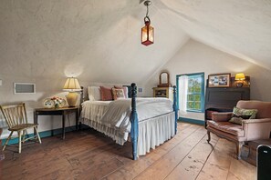 A bedroom with a wooden bed, a chair, a table with a lamp, and a window with curtains. - A 1787 home older than the country it stands in (Fredericksburg)