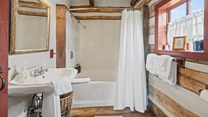 Een landelijke badkamer met houten inrichting, een witte badkuip, een wastafel met spiegel en een raam met gordijnen.