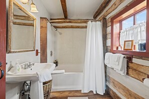 Un baño rústico con paredes de madera, una bañera blanca, un lavabo con espejo y una ventana con cortinas.