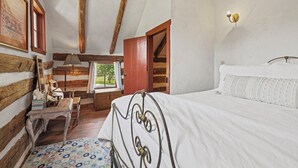 Ein Schlafzimmer mit Holzbalkendecke, einem Bett mit weißer Bettdecke, einem Holztisch, einem Stuhl, einer Lampe und einem Fenster mit Blick auf Grünflächen.