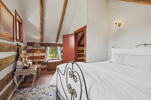 Une chambre avec un plafond en bois, un lit recouvert d’une courtepointe blanche, un bureau en bois, une chaise, une lampe et une fenêtre donnant sur un paysage verdoyant.