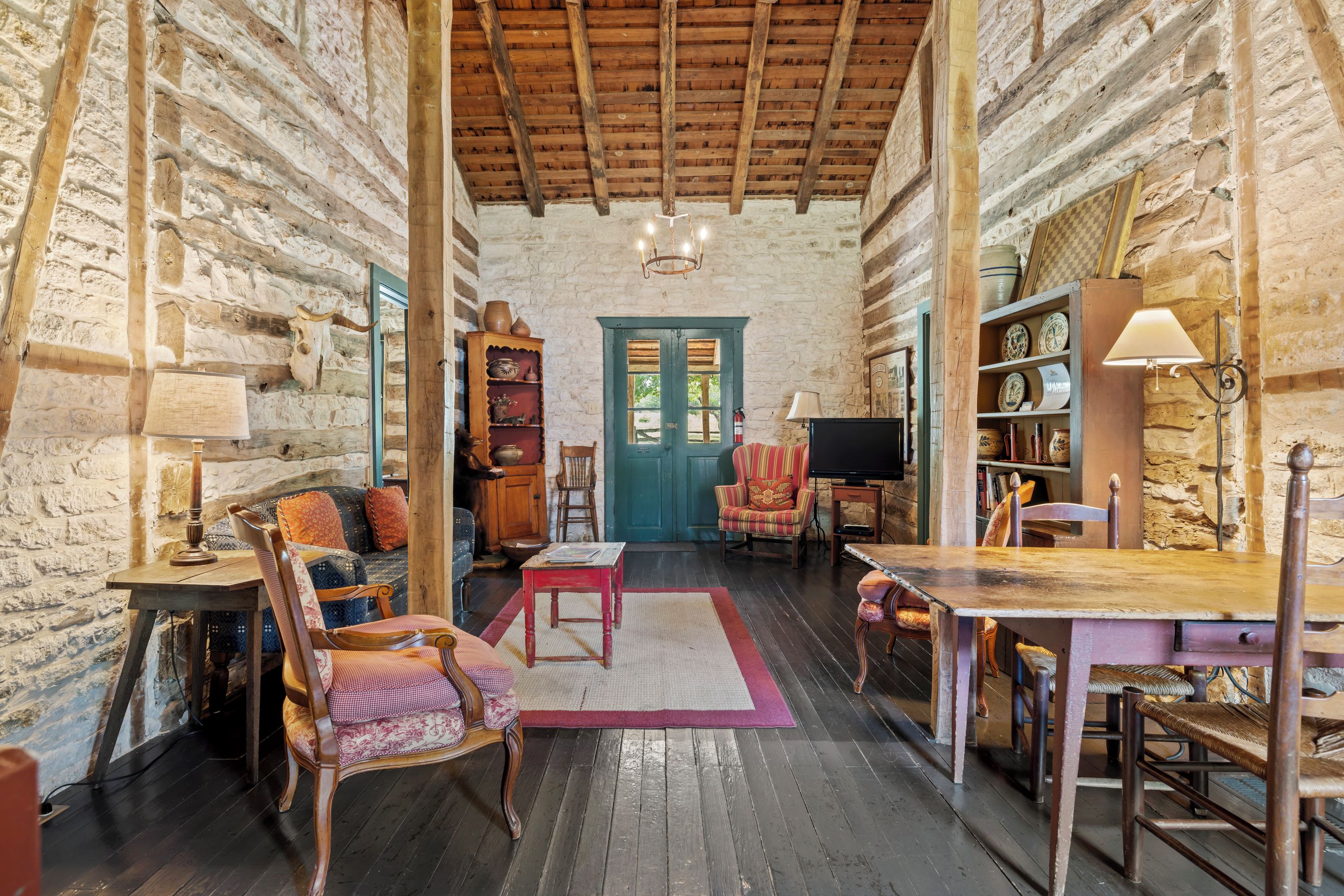 A rustic interior with exposed stone walls, wooden beams, a wooden table, a television, and a bookshelf.